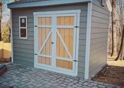 Completed garden shed with grey siding, white trim, and double wooden barn doors on stone paving.