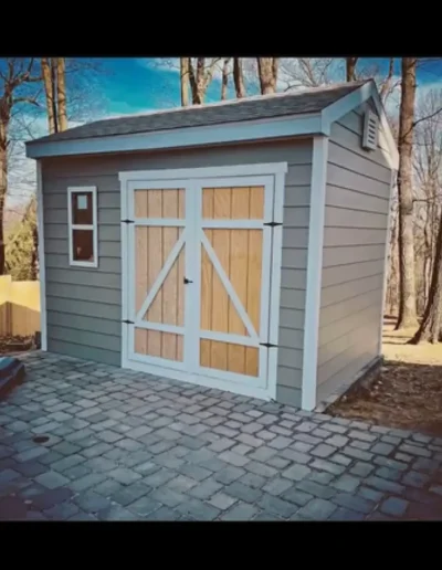 Completed garden shed with grey siding, white trim, and double wooden barn doors on stone paving.