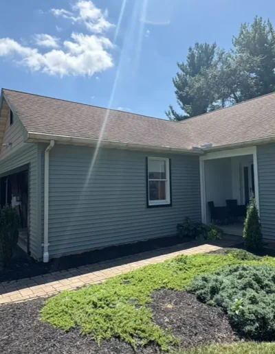 Front view of renovated home with new grey siding, brown shingle roof, and landscaped garden walkway on a sunny day.