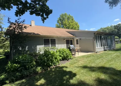 Back view of a renovated home with brown shingle roof, grey siding, patio area, and garden under clear blue sky.