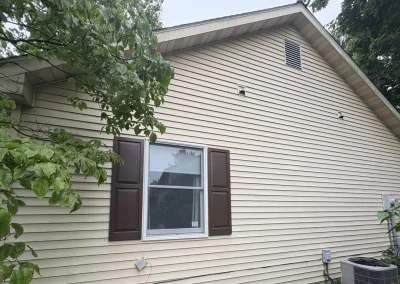 House exterior with beige vinyl siding, brown shutters, and trees surrounding the wall area.