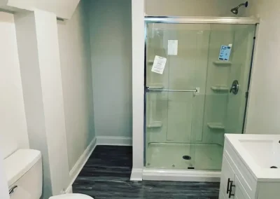 Small bathroom with glass shower enclosure, white vanity, toilet, and dark wood-style flooring.