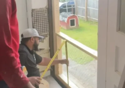 Two workers measuring wall opening for new window installation with outdoor backyard visible through the frame.