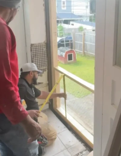 Two workers measuring wall opening for new window installation with outdoor backyard visible through the frame.