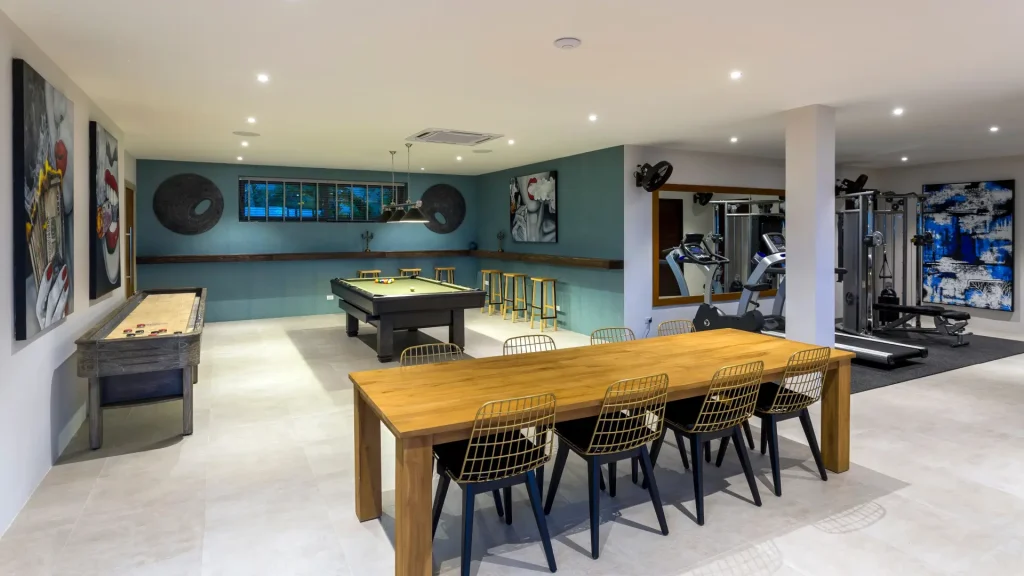 Modern basement entertainment and gym area featuring a pool table, exercise equipment, shuffleboard table, and wooden dining table with stylish chairs and wall art.
