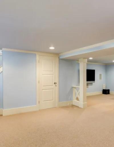 Spacious finished basement with light blue walls, beige carpet flooring, sectional sofa with red pillows, and a wall-mounted TV near the staircase.