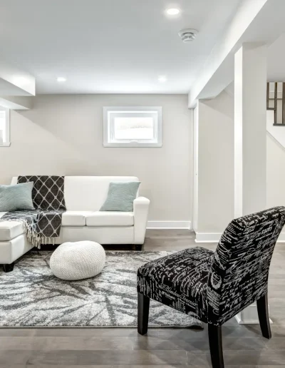 Modern finished basement with white sectional sofa, patterned rug, accent chairs, recessed lighting, and staircase with wooden steps and black railing.