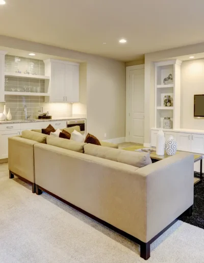 Stylish finished basement living room with beige sectional sofa, built-in TV unit, armchair, and small bar area with cabinets and lighting.