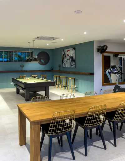 Modern basement entertainment and gym area featuring a pool table, exercise equipment, shuffleboard table, and wooden dining table with stylish chairs and wall art.