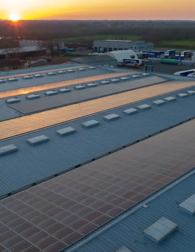 Industrial building rooftop with rows of solar panels installed on a metal roof, illuminated by the setting sun, surrounded by warehouses and open green fields.