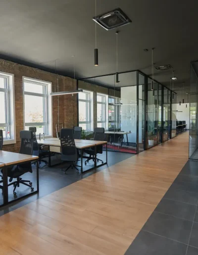 Modern industrial-style office with exposed brick walls, glass partitions, wooden flooring, ergonomic desks, and a lounge area with contemporary seating.