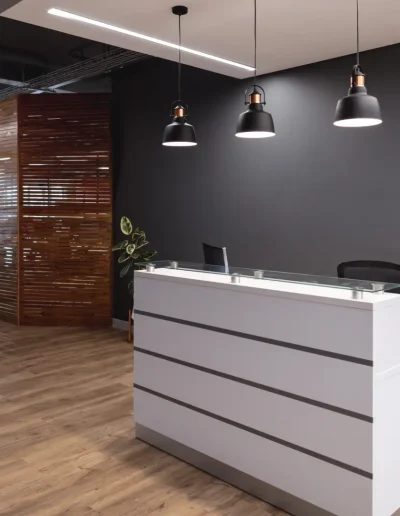 Modern office reception area with a sleek white desk, pendant lighting, dark accent wall, wooden flooring, and a stylish curved wooden partition in the background.