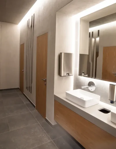 Modern restroom interior with sleek sinks, wooden doors, large mirrors, and ambient lighting, featuring minimalist décor and clean grey tile flooring.