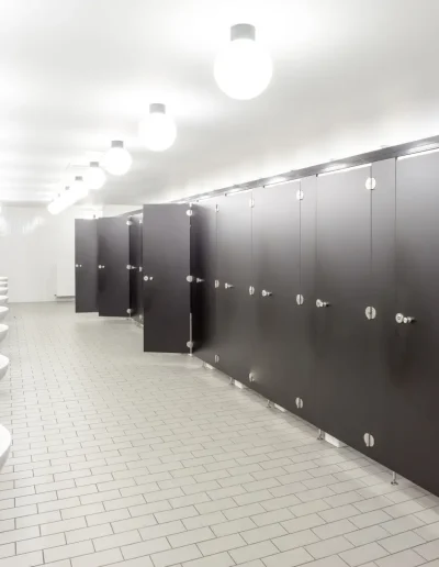 Bright public restroom with white tiled floors, multiple sinks along one wall, and black stall partitions under round ceiling lights for a clean, modern appearance.