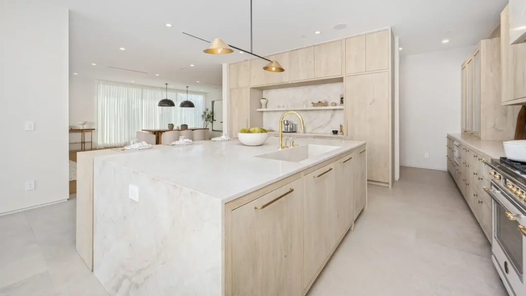 Luxury modern kitchen with light wood cabinetry, marble countertops, gold fixtures, and minimalist lighting, seamlessly connected to a bright open dining area.