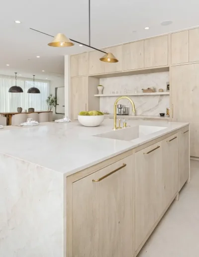 Luxury modern kitchen with light wood cabinetry, marble countertops, gold fixtures, and minimalist lighting, seamlessly connected to a bright open dining area.