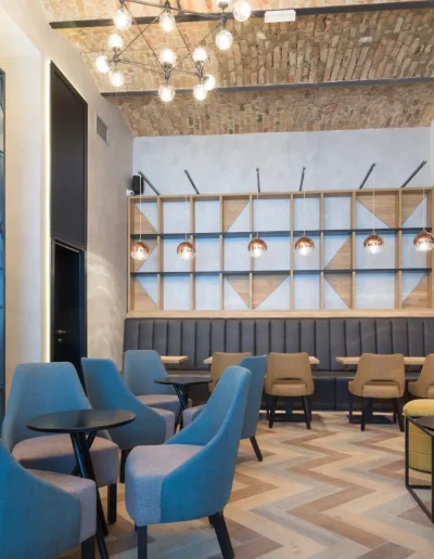 Modern café-style lounge with chevron wood flooring, exposed brick ceiling, elegant lighting, and a mix of blue and beige seating with contemporary décor.