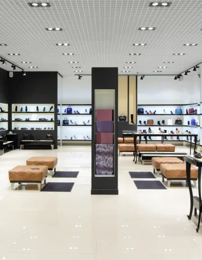 Modern retail shoe store interior with bright lighting, sleek shelving displays, tan seating benches, and glossy tiled flooring showcasing various footwear and accessories.