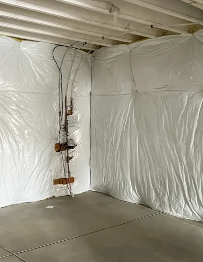 Unfinished basement with white plastic vapor barrier insulation on the walls, exposed ceiling beams, concrete flooring, and visible plumbing and electrical lines.