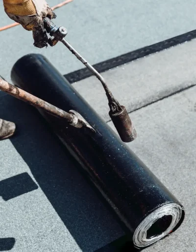 Workers installing a waterproof roofing membrane using a blowtorch, applying heat to seal and adhere the rolled material to the roof surface.