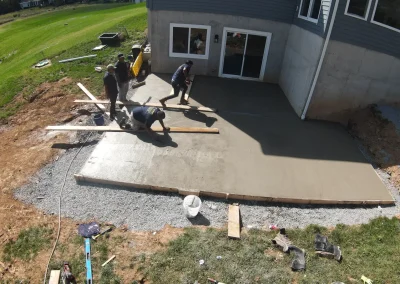 Aerial drone view of a concrete patio being poured and finished by a crew of workers behind a two-story gray residential home