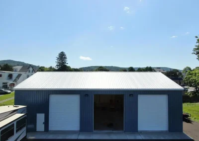 Completed metal workshop building with blue steel siding, white corrugated metal roof, and three garage bays set against a scenic hillside backdrop