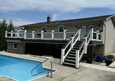 Full exterior view of a raised multi-level composite deck with white ornate railings, black balusters, staircase leading down to poolside concrete patio