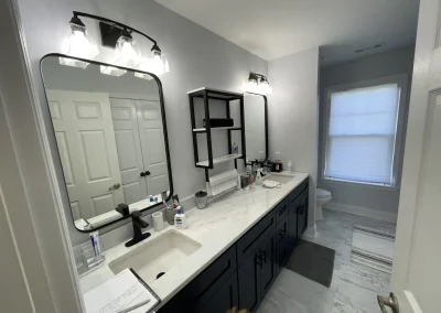 Wide-angle view of renovated master bathroom with double navy vanity, marble countertop, black-framed mirrors, industrial shelf unit, and marble-look tile flooring