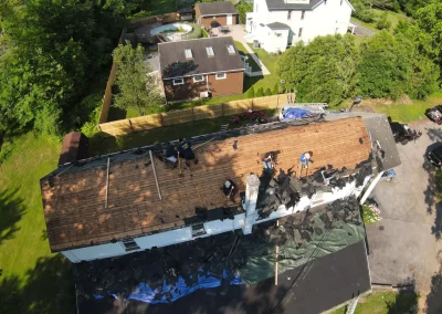 Aerial drone view of a residential roof tear-off in progress with workers removing old shingles, exposing bare wood decking on one side of the home