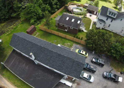 Aerial drone view of a completed new dark charcoal asphalt shingle roof on a residential home with surrounding neighborhood and lush green trees