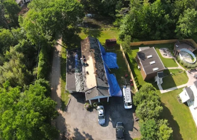 Aerial drone view of a residential roof replacement mid-project with workers removing old shingles, bare wood decking exposed, and blue tarps protecting the driveway and yard from debris