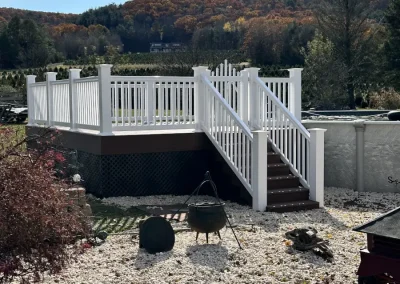 Completed pool access deck platform with white vinyl railings and matching staircase adjacent to an above-ground pool, set against an autumn foliage hillside backdrop