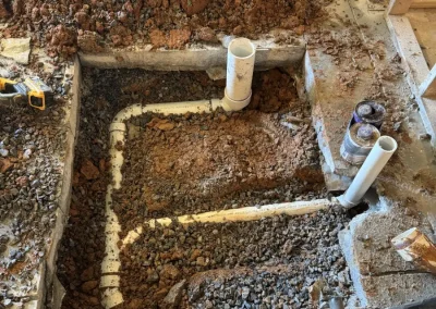 Underground plumbing rough-in showing excavated concrete floor with PVC drain pipes and fittings installed in gravel before backfill and concrete pour